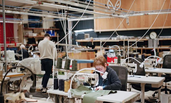 A Grandmaster sews denim at a workstation inside the Hiut Denim factory. Other team members work in the background beneath a wooden wall featuring the
